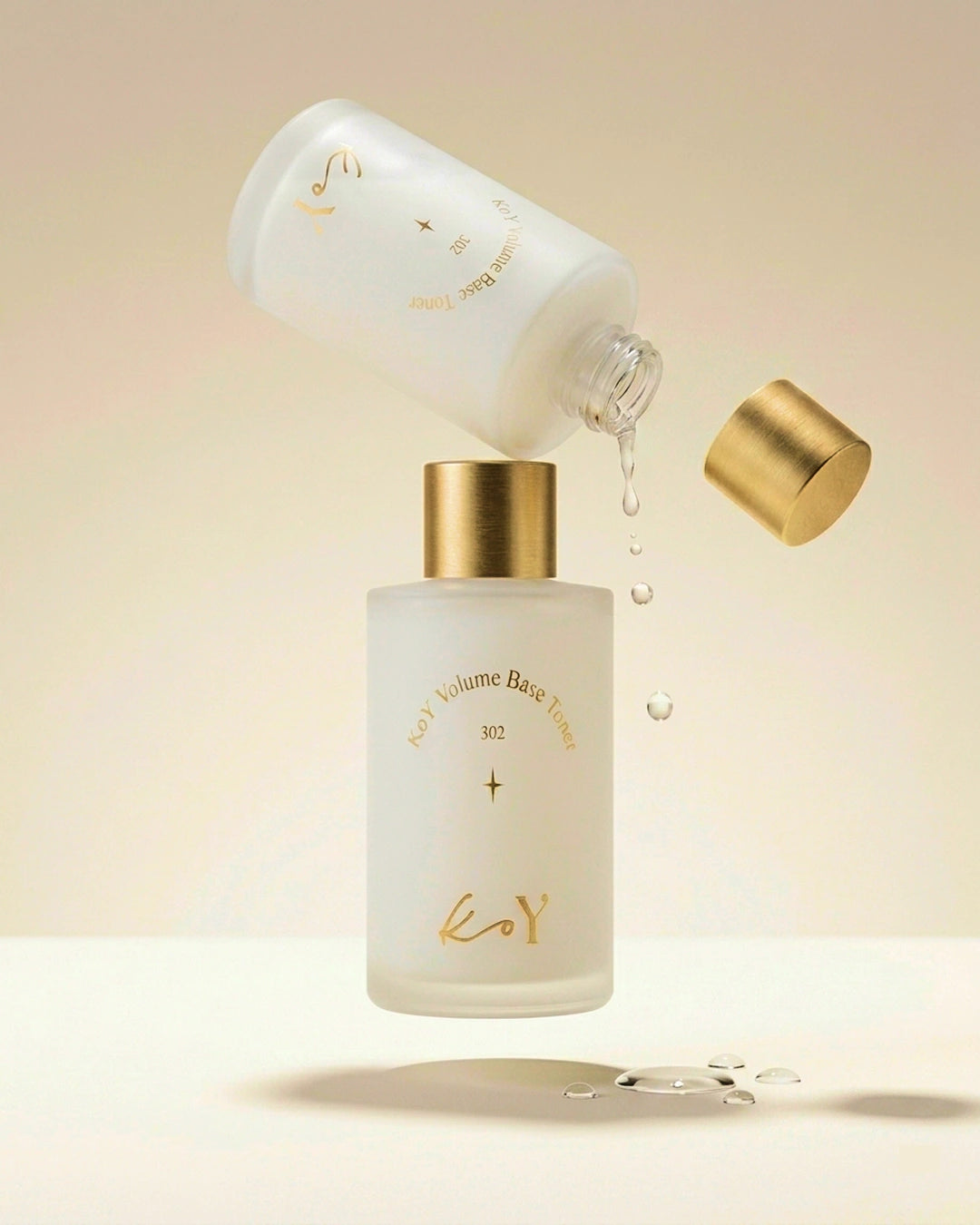 A frosted glass skincare bottle with a metallic gold cap, shown with a second tilted bottle pouring clear liquid into it. The bottles have gold lettering and are set against a warm beige background with a few floating water droplets.