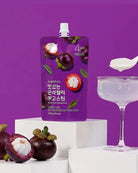 A purple stand-up pouch of sugartalk mangosteen-flavored sugar syrup stands on white display blocks, with real mangosteens and a glass coupe filled with clear syrup beside it against a solid purple background.