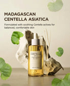 A bottle of SKIN1004 Madagascar Centella Asiatica Ampoule serum, featuring golden-yellow liquid in a clear bottle with a white dropper cap, styled with green leaves and natural stones on a neutral beige background.