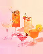 Five colorful cocktails in various glassware, including a coupe, highball, and martini glasses, arranged on a pink and peach gradient background with soft shadows.