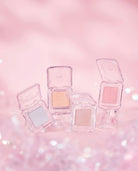 Four clear square eyeshadow compacts with open lids, displaying shimmering eyeshadow shades in silver, peach, pink, and champagne, arranged on a soft pink glittery background.