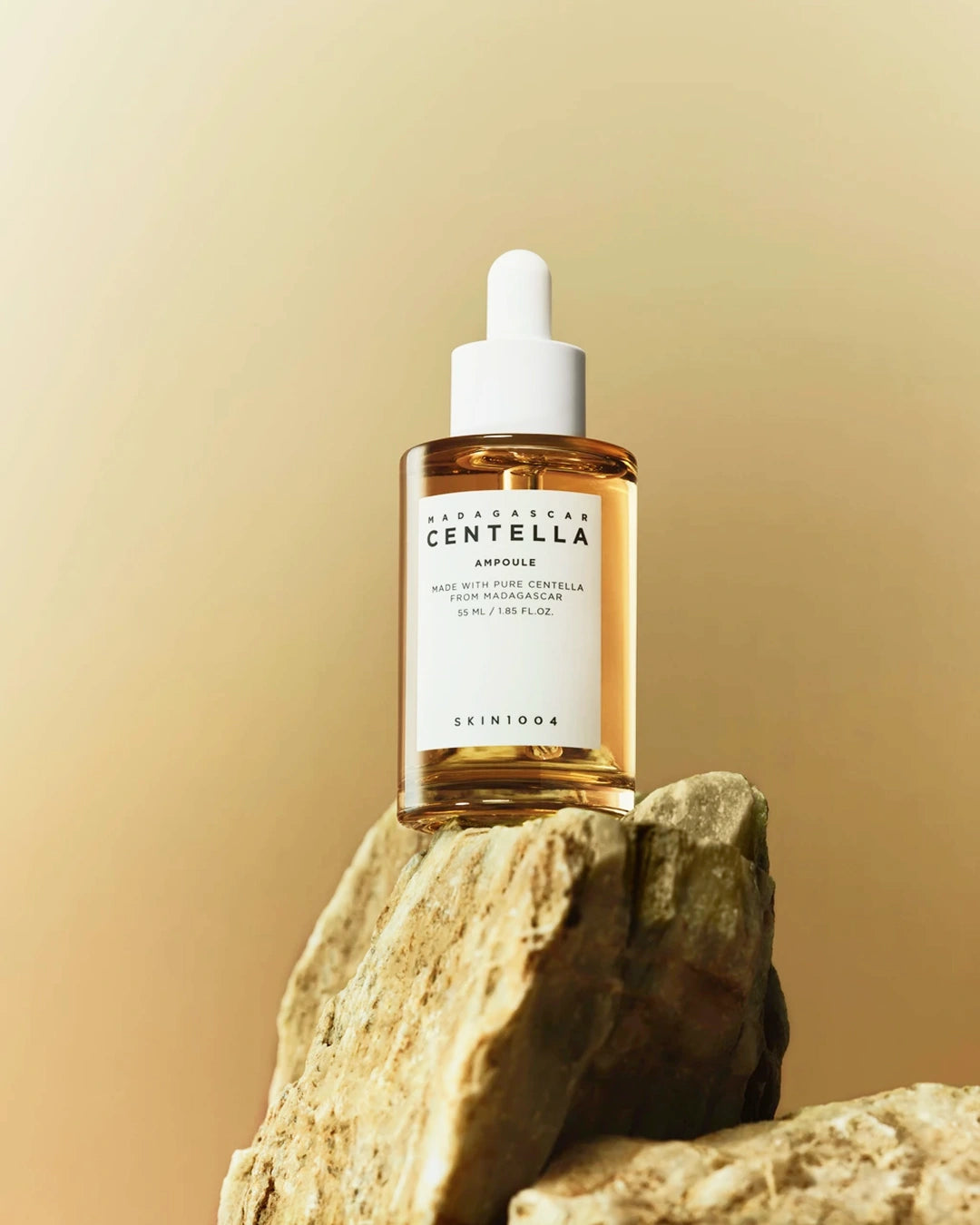 A bottle of SKIN1004 Madhavarsha Centella Ampoule serum, featuring a white dropper cap and a clear glass bottle filled with golden-amber liquid, displayed on a textured stone against a warm beige gradient background.