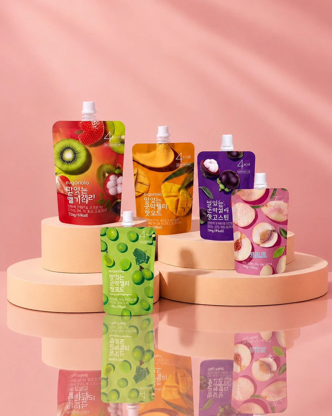 Five colorful pouches of fruit-flavored drink mix arranged on pastel display risers against a soft pink background. The pouches feature fruit illustrations including kiwi, strawberry, mango, blueberry, and peach, with Korean text on the front.