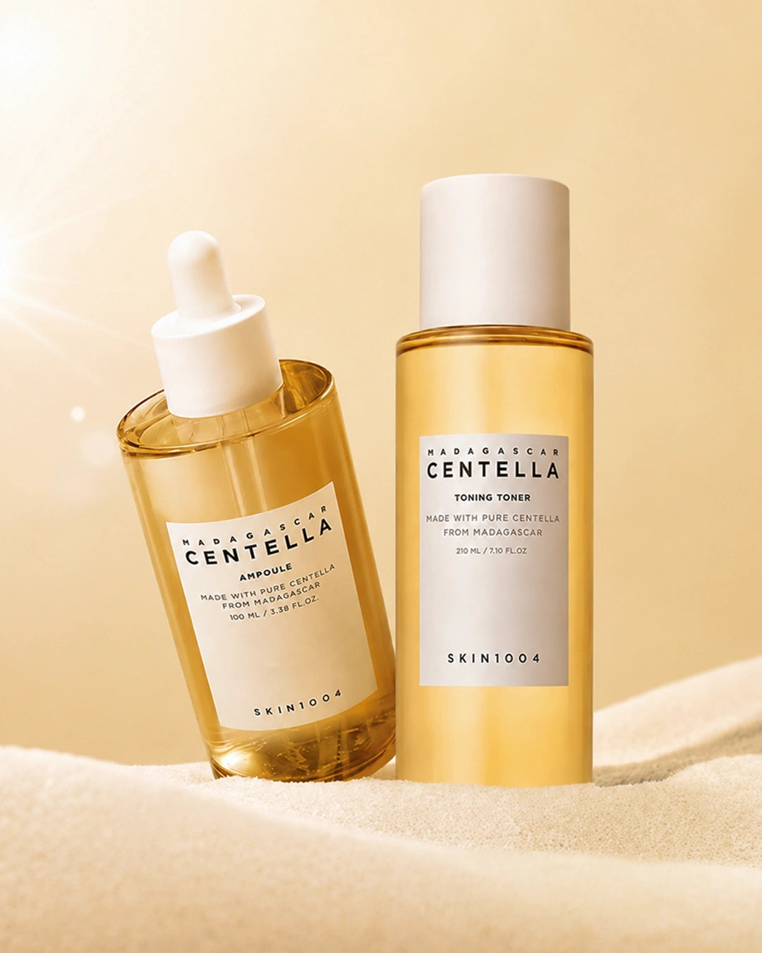 Two SKIN1004 Herbal Essence Centella skincare bottles on a soft beige fabric surface with a warm beige background. One bottle is a clear dropper bottle containing golden-yellow serum, and the other is a cylindrical toning tower bottle filled with pale yellow liquid.