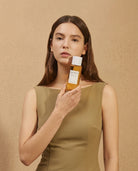 A woman in a sleeveless olive green dress holds a bottle of REVELA hair product against a warm beige textured background.