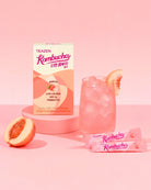 TEAZEN Rombucha Peach flavor probiotic drink packaging shown with a pink iced drink in a glass, two sachets, and peach halves on a soft pink studio background.