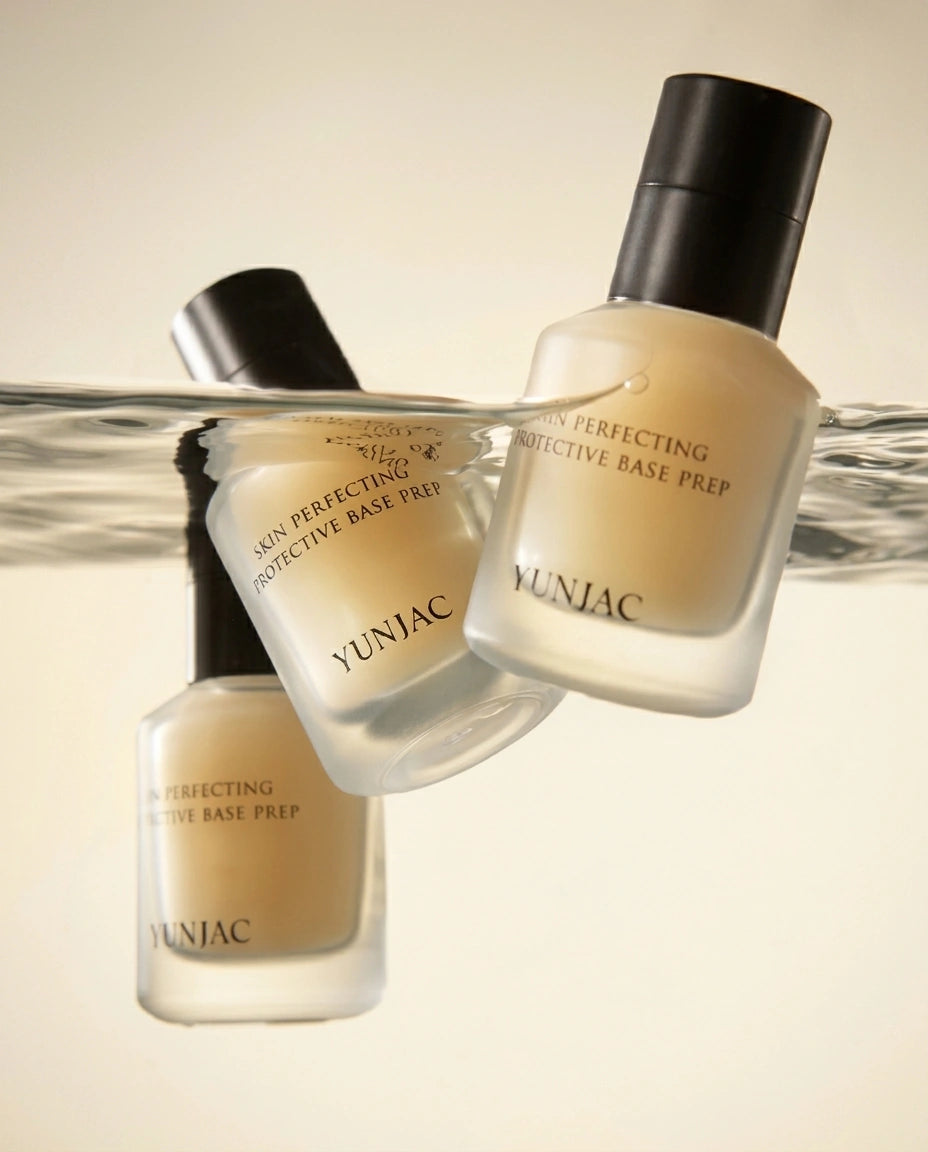 Three bottles of YUNJAC Perfecting Protective Base Prep makeup cream floating above a water surface, with one bottle reflected below. The bottles are frosted with a warm beige liquid and have black caps, set against a clean beige background.