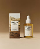 SKIN004 Centella Ampoule shown with its matching product box on a warm beige background. The box features a desert landscape print, while the clear glass bottle contains a golden-yellow liquid and has a white dropper cap.