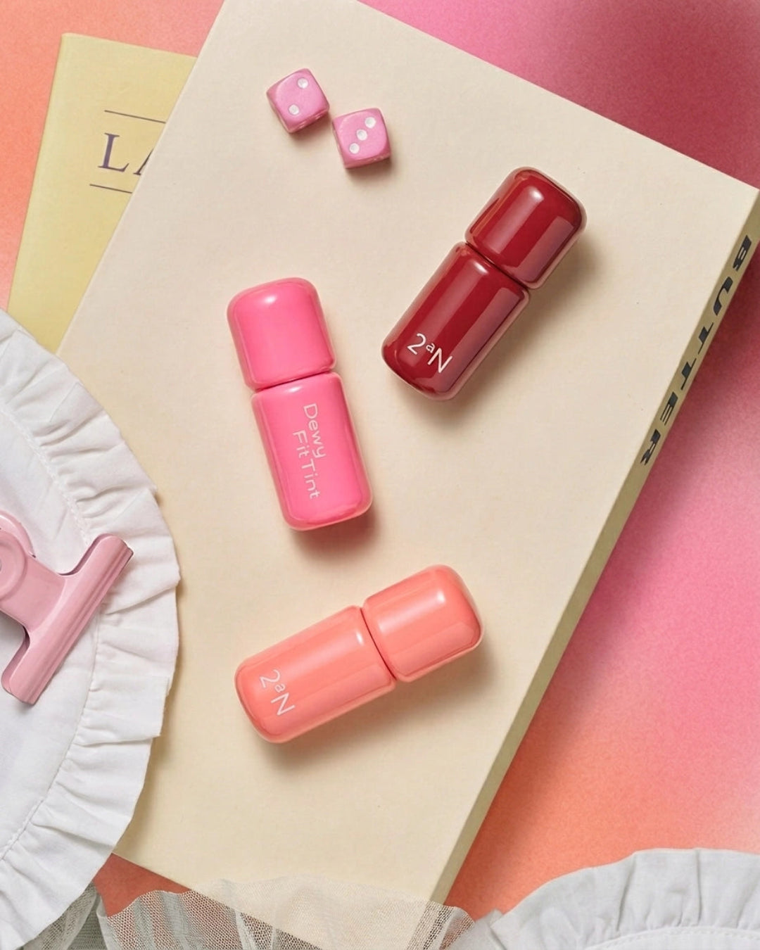Three small cylindrical cosmetic bottles in pink, coral, and deep red are arranged on a cream book with pink dice and a white ruffled fabric prop on a pink background.
