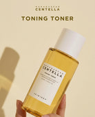 A hand holding a bottle of SKIN1004 Madagascar Centella Toning Toner, which has a translucent golden-yellow liquid and a white cap, set against a warm beige background with text overlays.