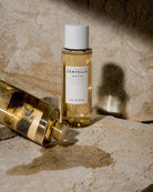 A bottle of SKIN 1004 Centella Tanse facial toner stands upright on a stone surface, with another bottle lying on its side nearby. The product is shown in warm golden liquid against a textured beige stone background with dramatic shadows.