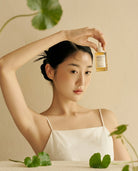 A woman in a white camisole holds a small amber bottle above her head against a warm beige wall with green leaves around her.