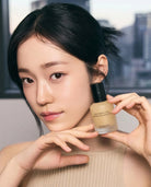A woman holds a bottle of Finiac Sun Protecting Moistive Base Paint against her face in a close-up shot. The bottle is a frosted beige color with a black cap, and the background is a soft-focus gray cityscape.