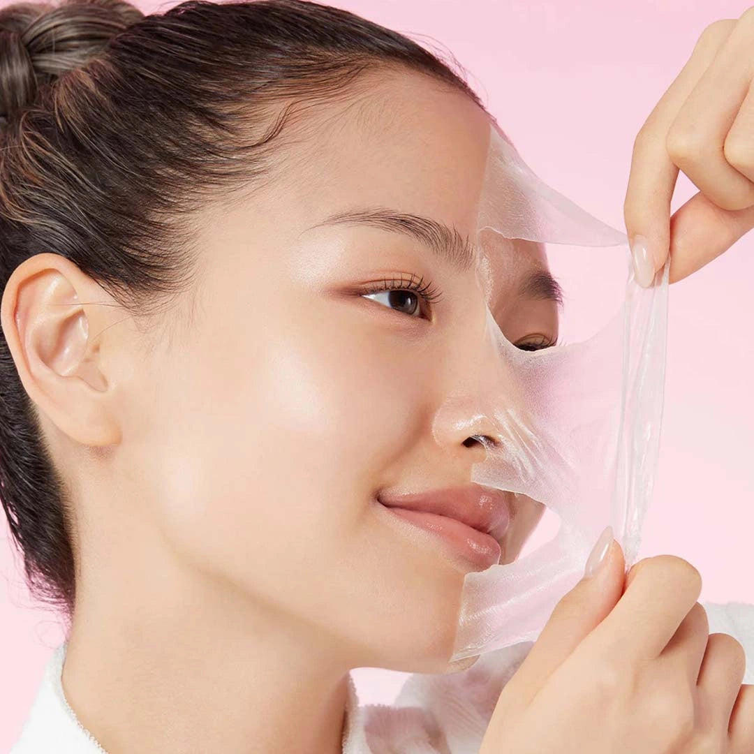 Woman applying a clear sheet mask to her face against a pink background