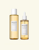 Two De'Nique skincare bottles on a light cream background: a larger clear bottle with pale golden liquid and a white cap, and a smaller dropper bottle with the same pale golden liquid and a white label.